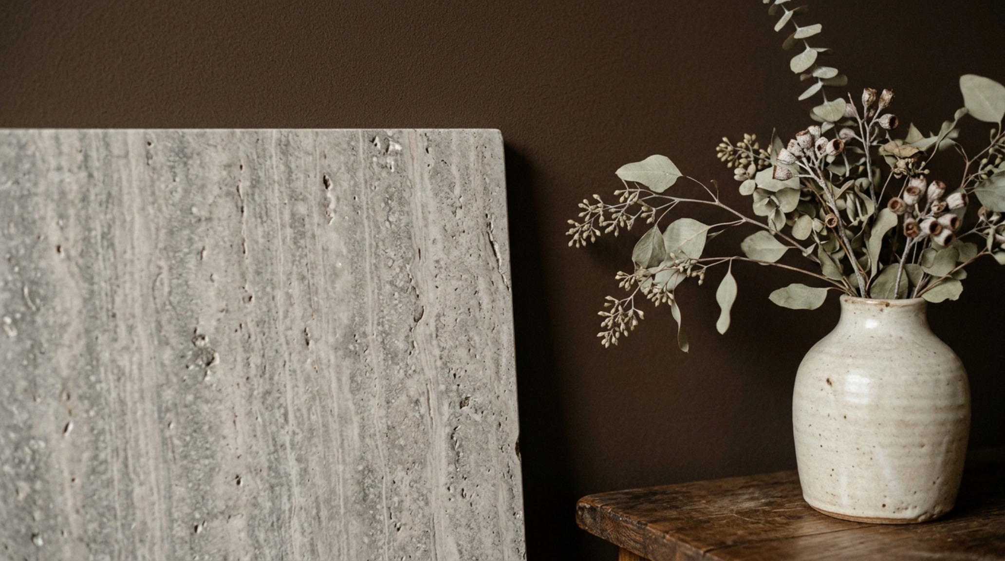 Close-up of tactile materials: a honed silver travertine surface against a textured brown limewash wall with 2700K warm ambient lighting.