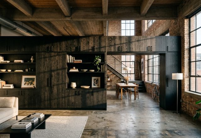 A wide-angle view of a modern black wood industrial loft with a custom floor-to-ceiling blackened oak storage unit acting as a room divider, featuring clean lines and 36-inch architectural walkways.