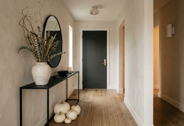 A sleek, narrow entryway featuring a minimalist black console table with tall architectural branches and white Casper pumpkins under soft 2700K lighting.