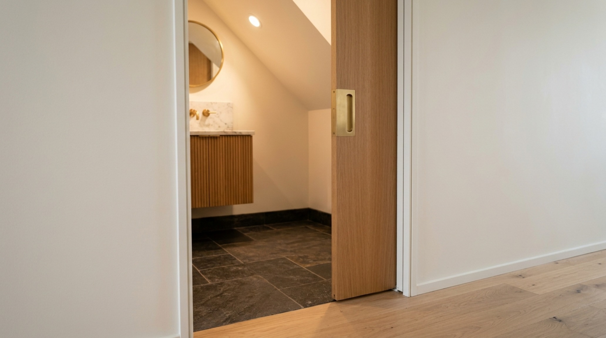 Detail view of a seamless pocket door entry into an under-stairs bathroom, showcasing the transition from a light oak hallway to a moody, slate-gray interior with a brushed brass handle. Detail view of a seamless pocket door entry into an under-stairs bathroom, showcasing the transition from a light oak hallway to a moody, slate-gray interior with a brushed brass handle.