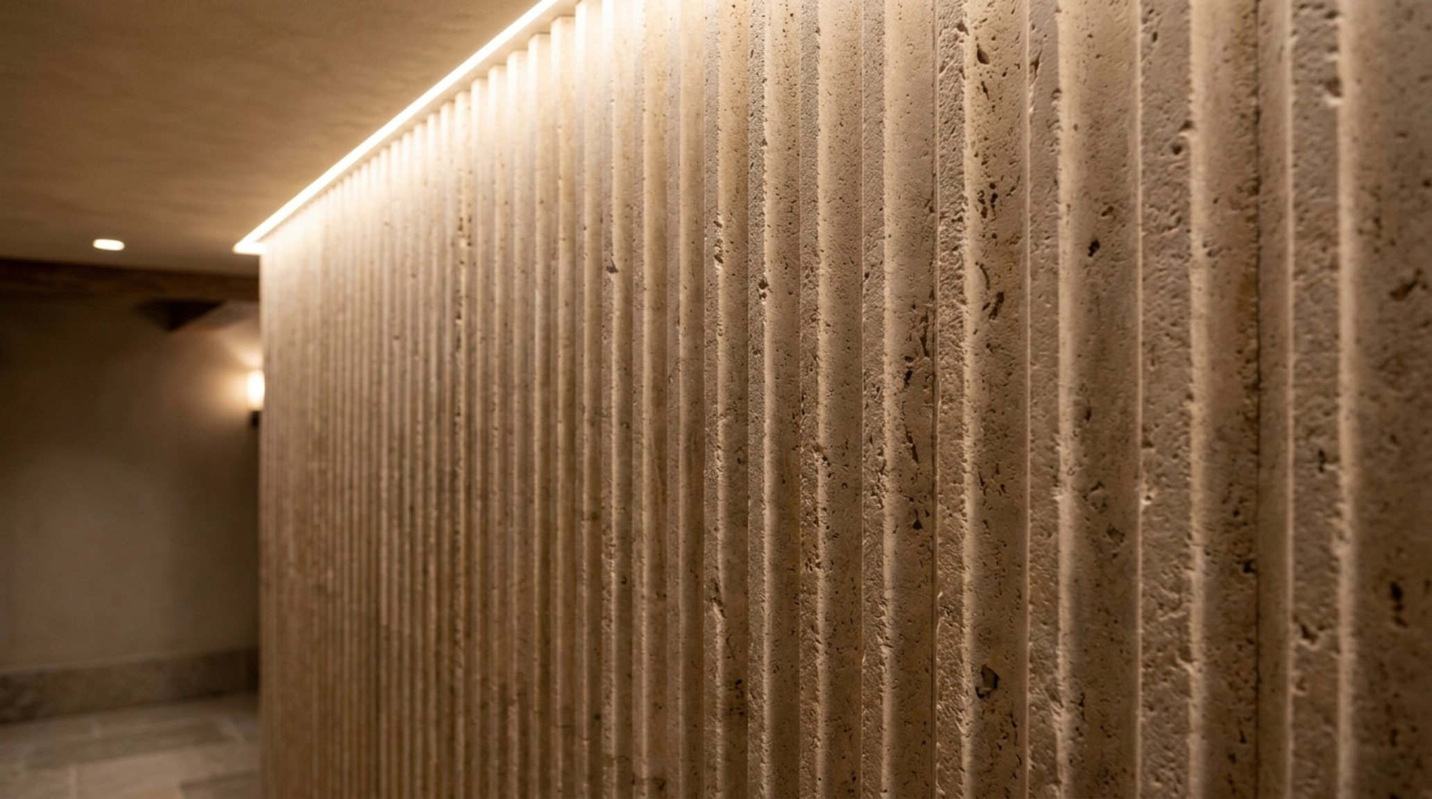 Soft cove lighting at 2700K washing over a honed travertine wall, emphasizing tactile grounding and the movement toward artisanal ceramic finishes in luxury homes.