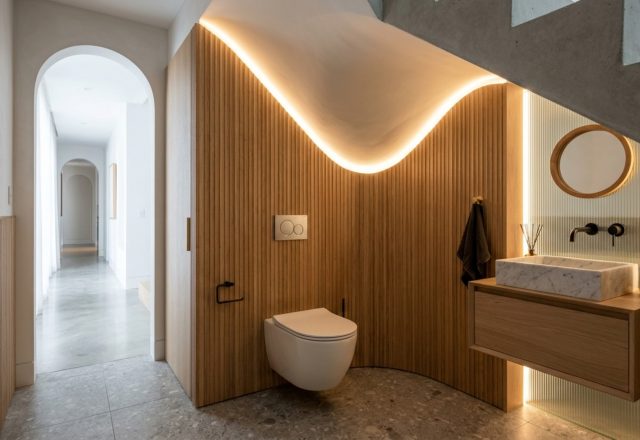Sophisticated powder room under stairs featuring a wall-hung Geberit toilet, vertical fluted oak wall panels, and soft warm cove lighting integrated into the sloping ceiling line.
