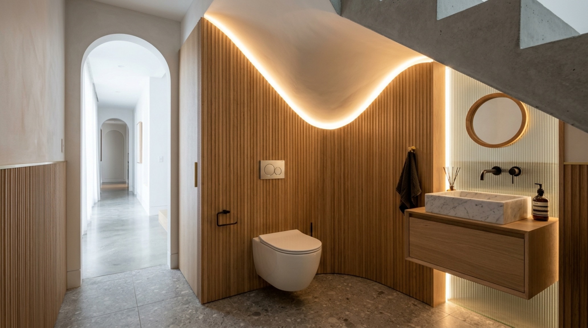 Sophisticated powder room under stairs featuring a wall-hung Geberit toilet, vertical fluted oak wall panels, and soft warm cove lighting integrated into the sloping ceiling line. Sophisticated powder room under stairs featuring a wall-hung Geberit toilet, vertical fluted oak wall panels, and soft warm cove lighting integrated into the sloping ceiling line.