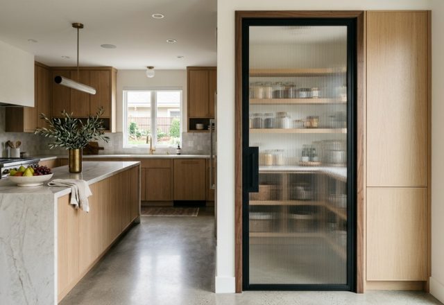 A narrow hallway view of a stylish pantry door with fluted glass and black steel frames, showing the architectural sightline into a bright kitchen.