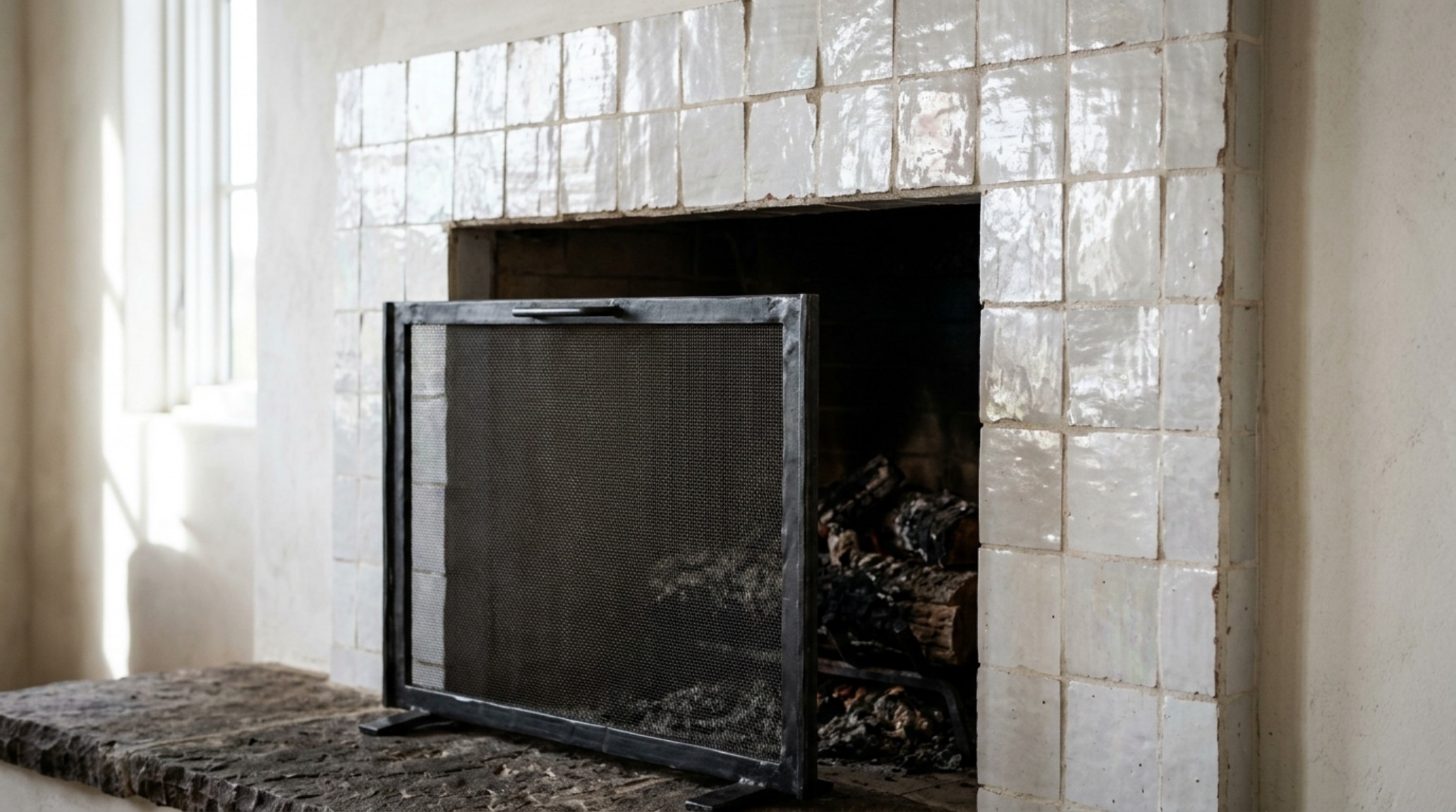 Detailed shot of Zellige tile fireplace surround in weathered white, showing high-gloss texture and irregular edges paired with a blackened steel fire screen.