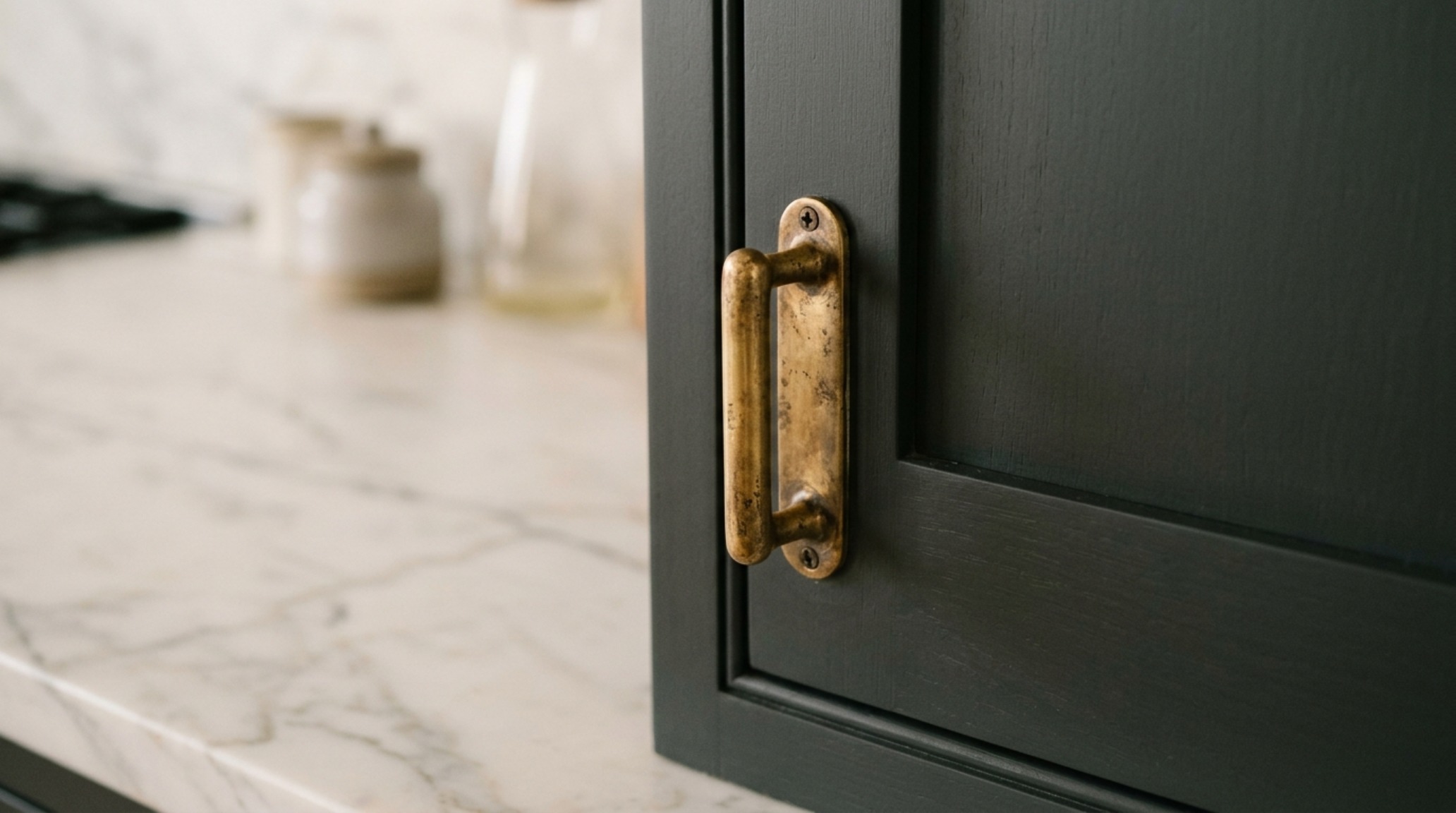 Close-up of unlacquered brass handles on matte charcoal cabinetry next to a honed white marble countertop with intricate honey-gold veining. Close-up of unlacquered brass handles on matte charcoal cabinetry next to a honed white marble countertop with intricate honey-gold veining.