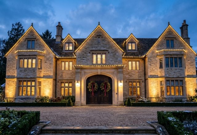 outdoor holiday house photography of a symmetrical limestone manor with warm LED lighting along the roofline and dual cypress wreaths on the entry doors