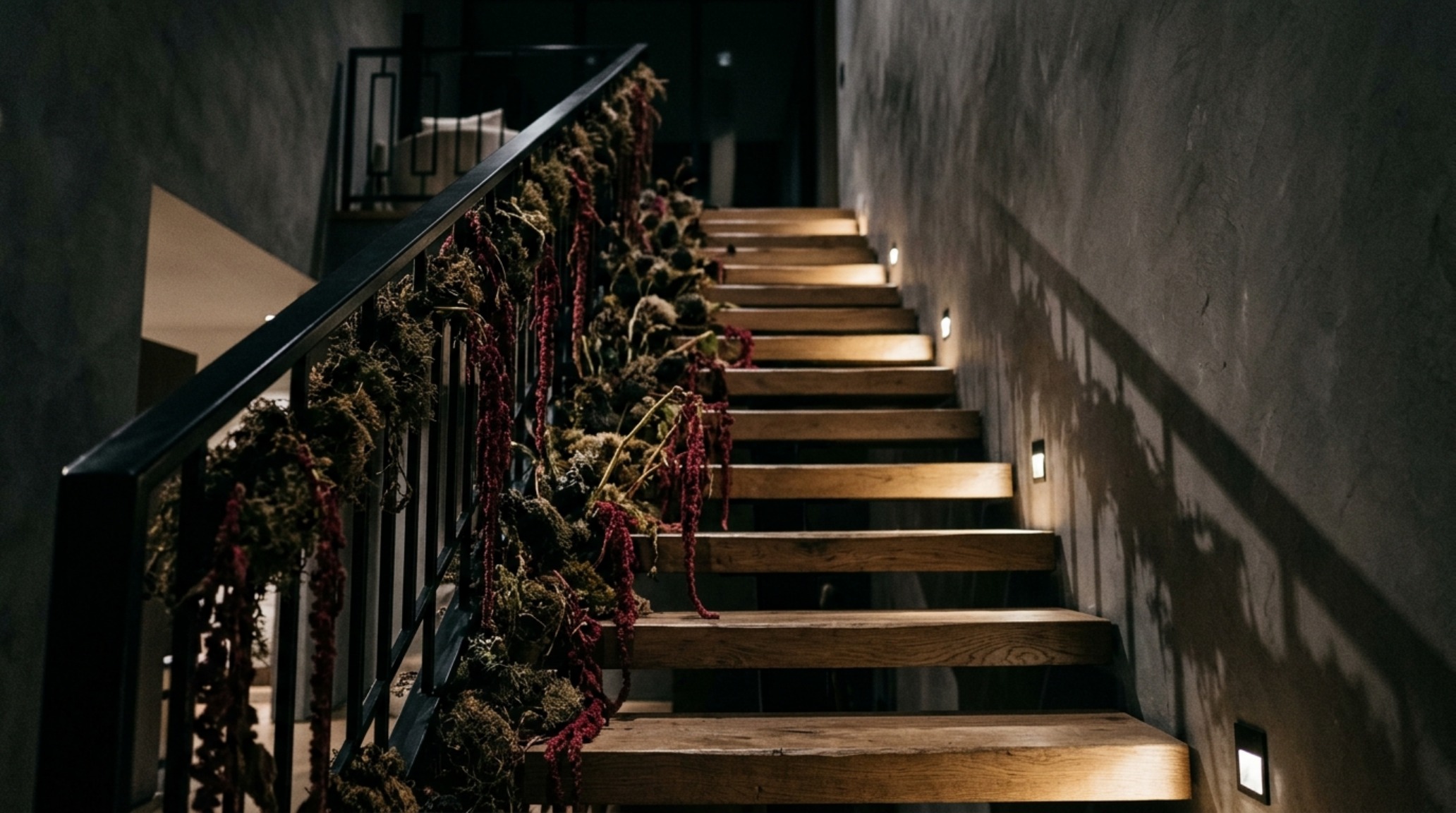 Moody seasonal lighting techniques used on a sculptural white oak staircase with trailing dark eucalyptus and low-angle amber uplighting.