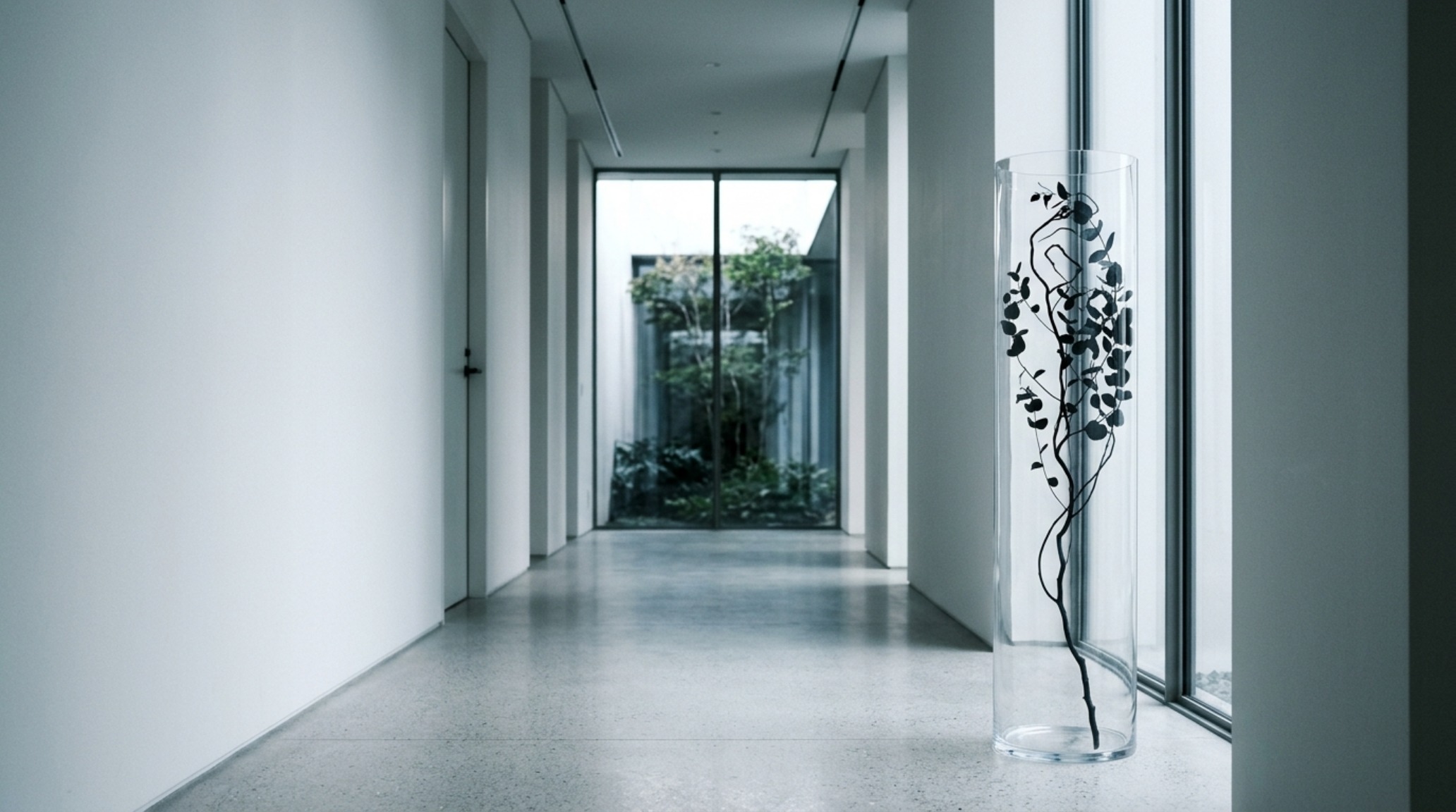 Minimalist halloween home decor focusing on negative space and geometry, with a floor-to-ceiling glass cylinder containing a single black-painted eucalyptus branch. Minimalist halloween home decor focusing on negative space and geometry, with a floor-to-ceiling glass cylinder containing a single black-painted eucalyptus branch.
