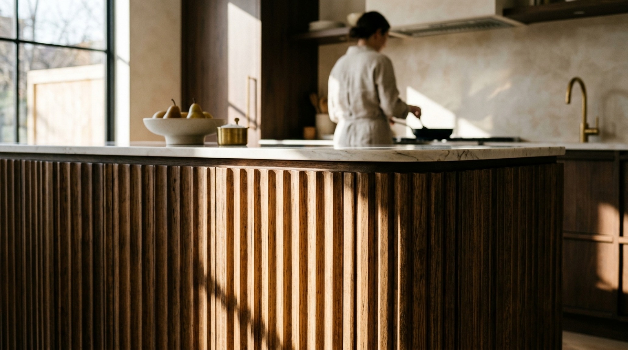 Fluted wood joinery on a floor-to-ceiling kitchen island crafted from white oak, catching natural morning sunlight through a large window. Fluted wood joinery on a floor-to-ceiling kitchen island crafted from white oak, catching natural morning sunlight through a large window.