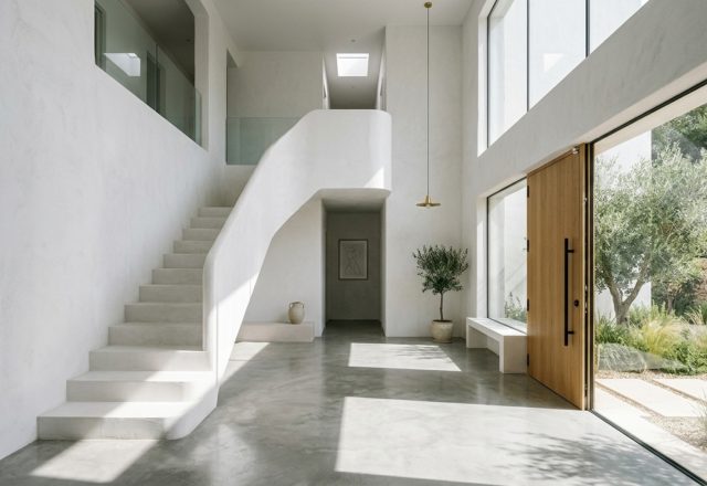 A soaring minimalist entryway featuring double-height white walls, a sculptural floating staircase, and a large pivot door, demonstrating architectural proportions in a minimalist white house.