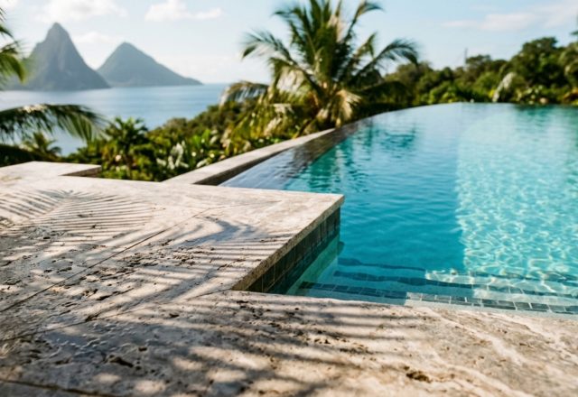 luxury st lucia infinity pool hotel textures featuring honed silver travertine pool coping meeting a matte volcanic stone wall under soft morning sunlight