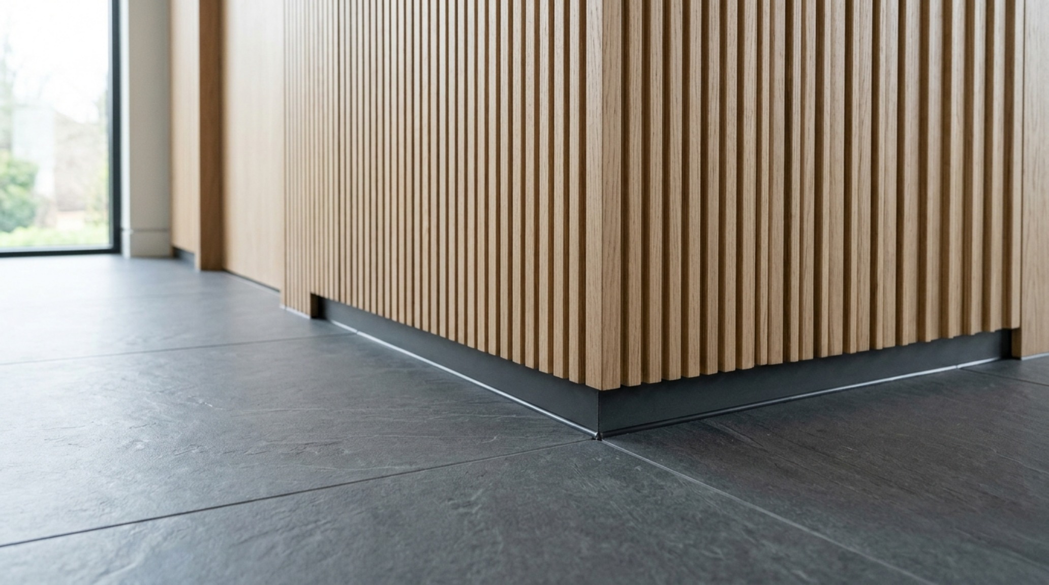 Architectural junction of matte slate floor tiles and fluted white oak cabinetry with a minimal 1/8-inch recessed shadow gap skirting.