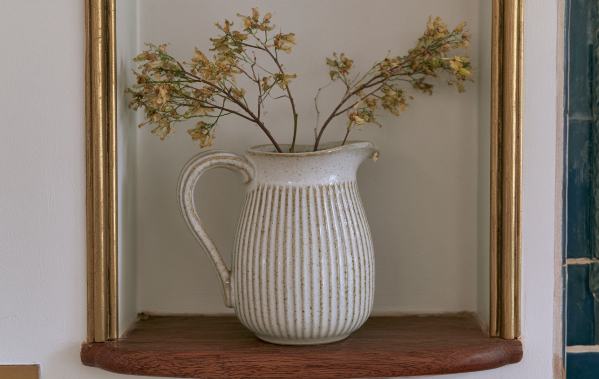 Minimalist alcove decor featuring a ribbed white ceramic pitcher with dried botanical stems on a wooden shelf.
