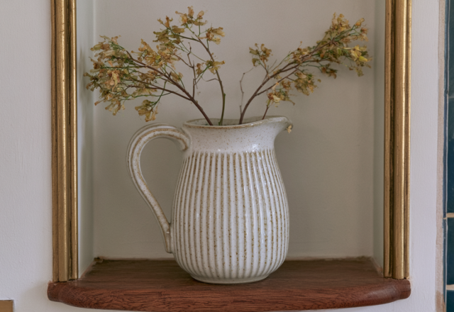 Minimalist alcove decor featuring a ribbed white ceramic pitcher with dried botanical stems on a wooden shelf.