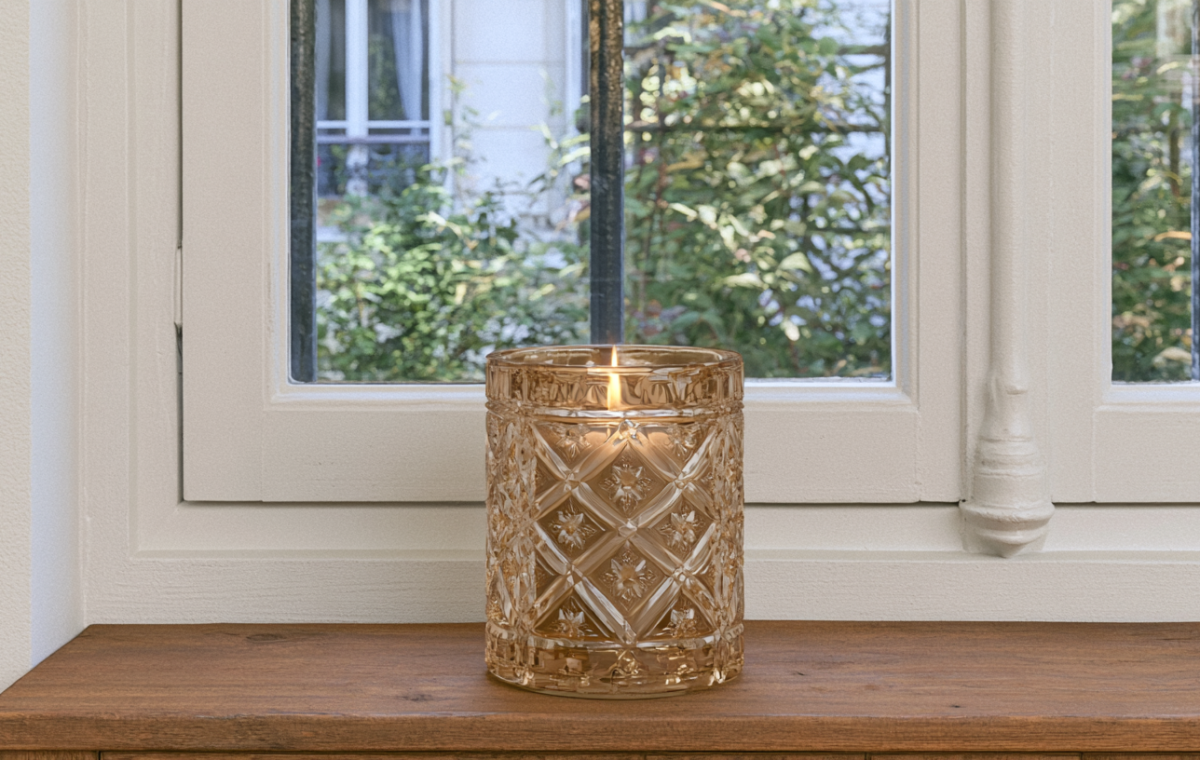An amber glass candle with a lit flame sits on a wooden windowsill overlooking green foliage.
