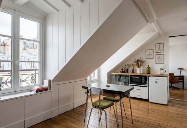 Bright attic apartment interior featuring slanted whitewashed walls, hardwood floors, and a compact dining table near a large window.