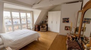 Bright attic bedroom design featuring sloped ceiling beams, parquet flooring, white wardrobes, and a dormer window overlooking city rooftops.