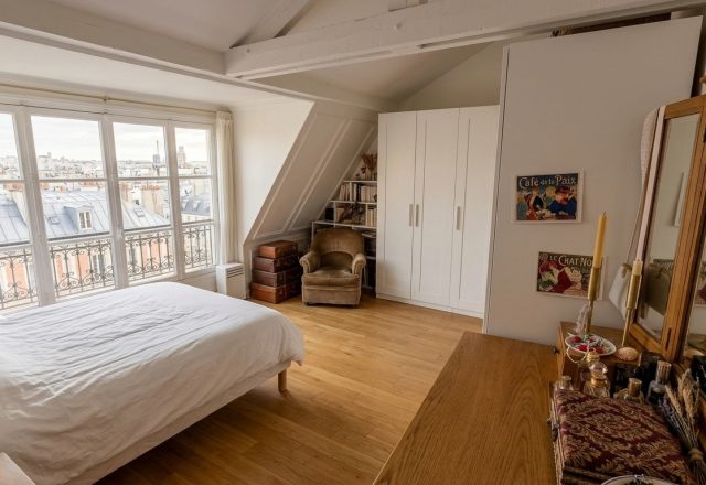 Bright attic bedroom design featuring sloped ceiling beams, parquet flooring, white wardrobes, and a dormer window overlooking city rooftops.