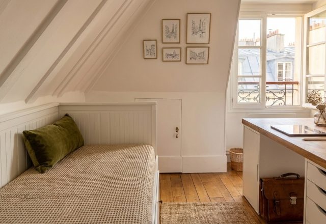 Cozy attic guest bedroom with sloped white rafters, daybed, and a wooden desk near a sunny window.