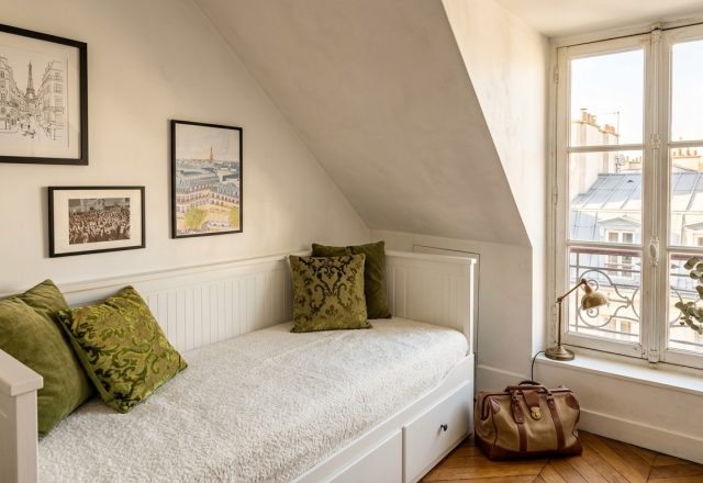 Cozy attic guest bedroom with white daybed, olive green pillows, and slanted walls under natural window light.
