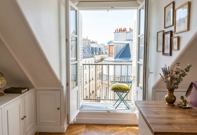 Bright attic home office ideas featuring sloped white walls, built-in storage, and open French doors leading to a balcony.