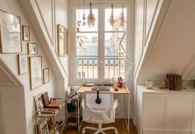 Cozy attic home office setup with slanted white walls, wooden desk under windows, and ergonomic chair.
