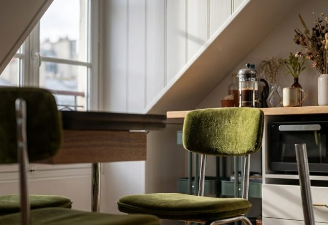 Cozy attic kitchenette design with sloped ceilings, green velvet chairs, and a coffee station.