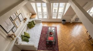 Bright attic living room with sloped ceilings, a white daybed, chevron wood floors, and twin dormer windows.
