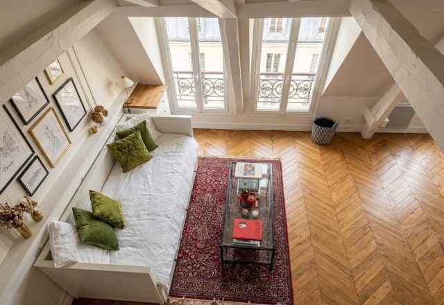 Bright attic living room with sloped ceilings, a white daybed, chevron wood floors, and twin dormer windows.