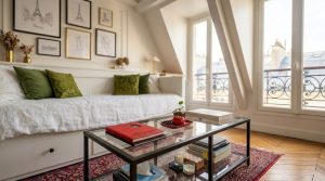 Cozy attic living room featuring a white daybed, glass coffee table, and slanted ceilings under tall windows.