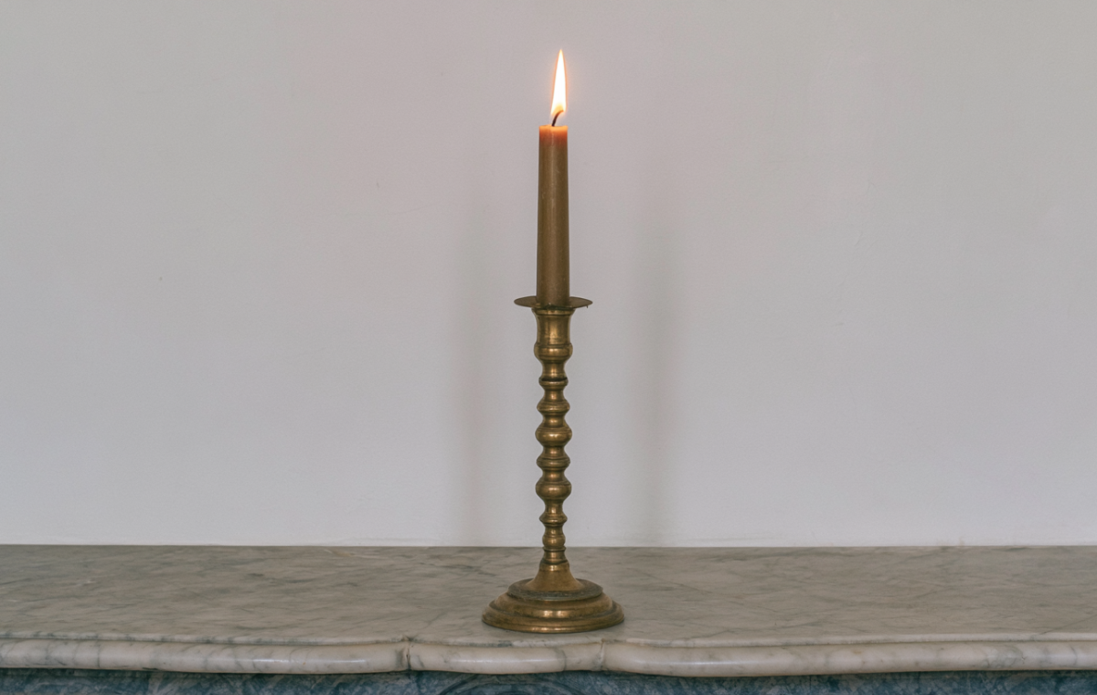 A slender lit brass candlestick holder sitting centered on an antique marble fireplace mantel against a white wall.