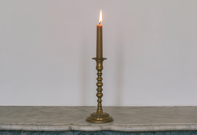 A slender lit brass candlestick holder sitting centered on an antique marble fireplace mantel against a white wall.
