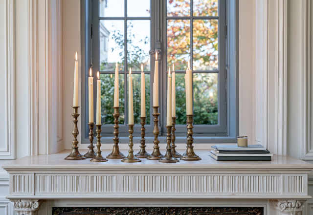 A curated collection of brass candlesticks mantle decor with lit taper candles arranged on a classic stone fireplace ledge.