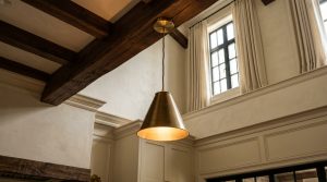 A modern brass pendant light hanging from a vaulted ceiling with dark exposed wood beams in a bright transitional kitchen.