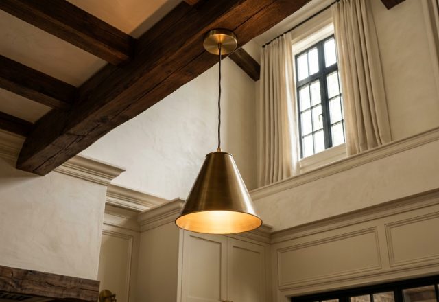 A modern brass pendant light hanging from a vaulted ceiling with dark exposed wood beams in a bright transitional kitchen.
