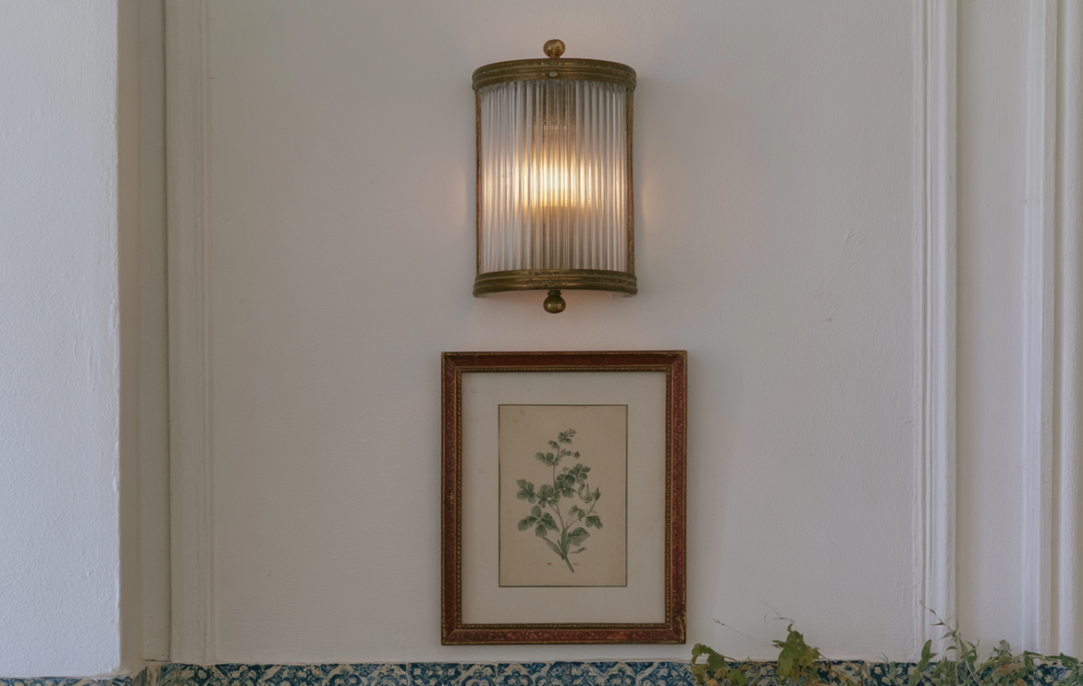 A warm brass wall sconce lighting fixture mounted above a framed botanical print in a bright hallway.
