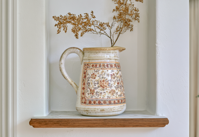 A rustic patterned ceramic pitcher filled with dried botanicals sitting inside a white built-in wall niche on a wood shelf.