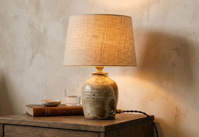 A textured ceramic bedside lamp with a linen shade glowing softly on a wooden nightstand in a neutral room.