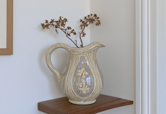 A rustic ceramic jug vase with dried stems on a wooden floating shelf against a white wall.