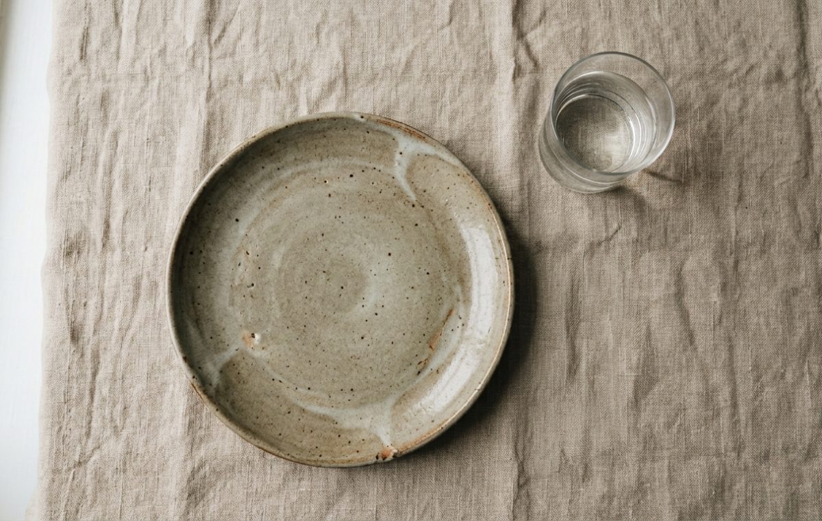 A minimalist stoneware ceramic plate set with a glass of water on a neutral linen tablecloth.
