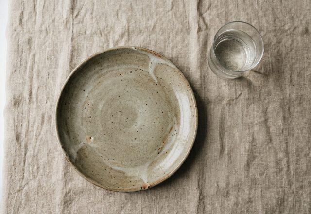A minimalist stoneware ceramic plate set with a glass of water on a neutral linen tablecloth.