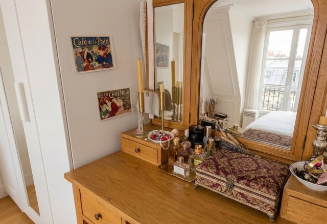 A warm wooden vanity setup featuring a triple mirror, vintage art prints, and organized perfume bottles.