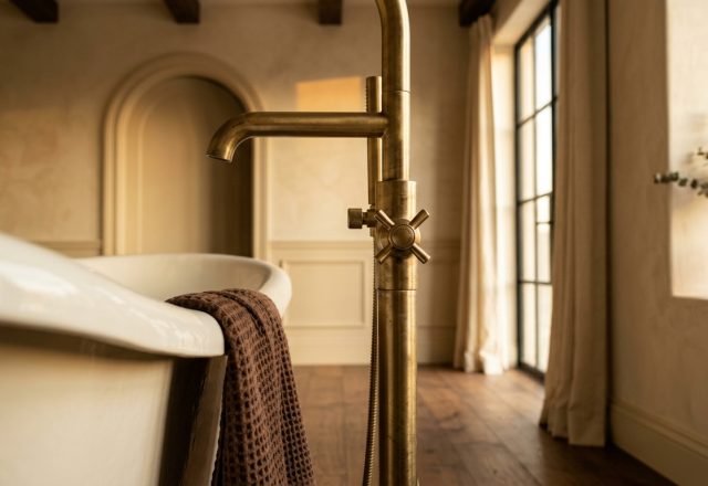 Close up of a freestanding brass bathtub faucet next to a deep soaking tub in a warm rustic bathroom.