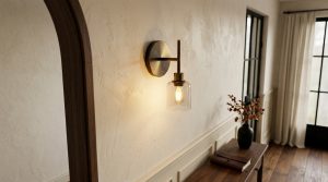 A stylish brass hallway wall sconce with a clear glass shade illuminating a textured plaster corridor wall.