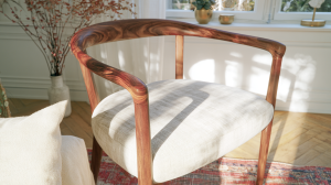 A sleek wooden armchair with a neutral upholstered seat sitting in a sunlit living room with herringbone floors.
