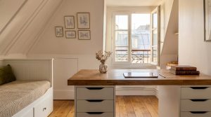 Bright attic home office setup featuring a wooden desk beneath sloped white ceilings and a window view.