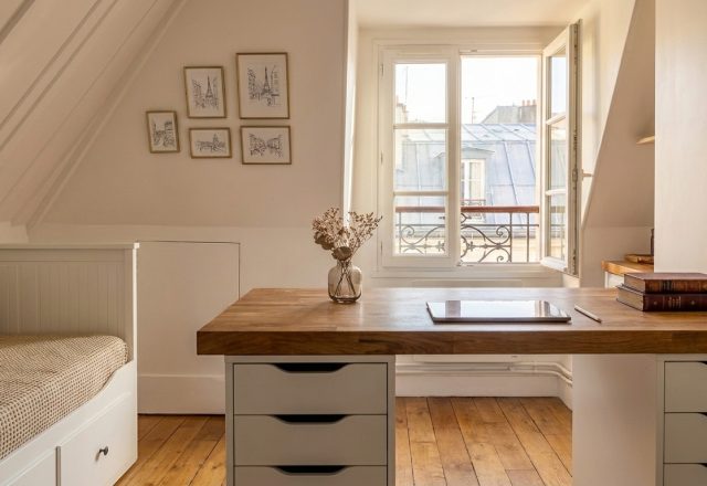 Bright attic home office setup featuring a wooden desk beneath sloped white ceilings and a window view.