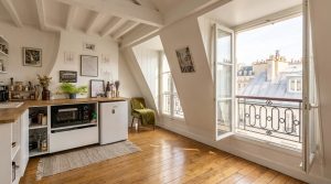Bright attic kitchen featuring sloped ceilings, wooden countertops, and a view of Parisian rooftops through casement windows.