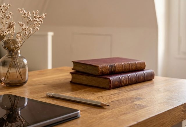 A minimalist home office setup featuring a wooden desk, antique books, dried flowers in a glass vase, and a tablet.